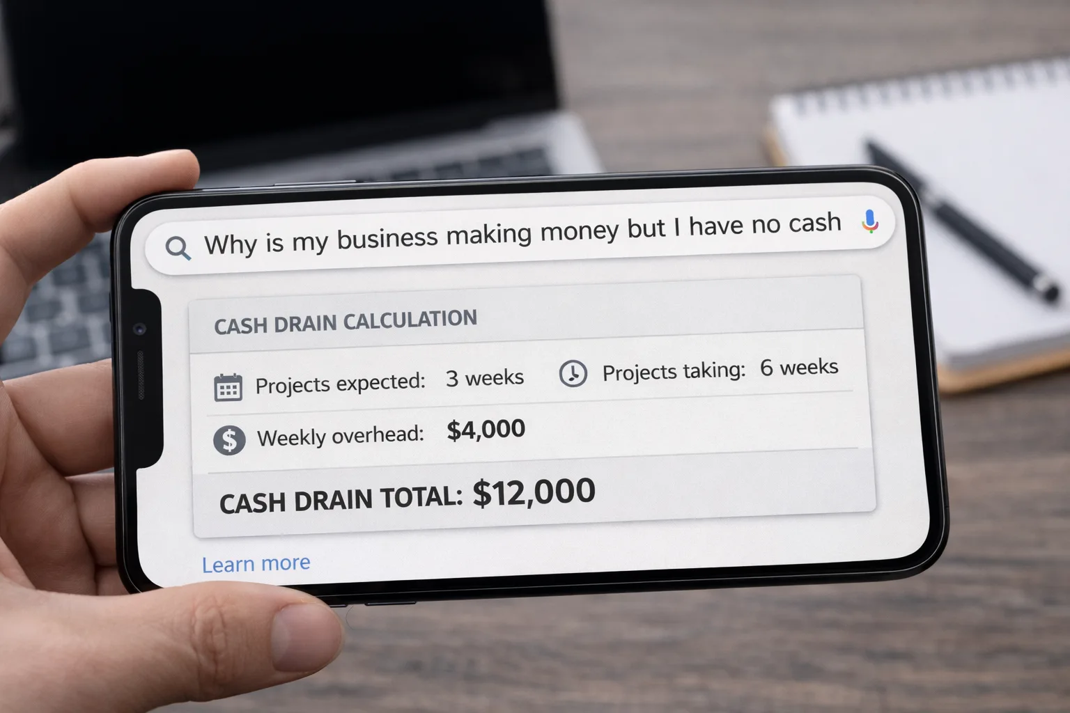 Smartphone in landscape mode showing a Google search for “why is my business making money but I have no cash” with a cash drain calculation displayed on screen, held over a desk workspace with a laptop and notebook in the background.